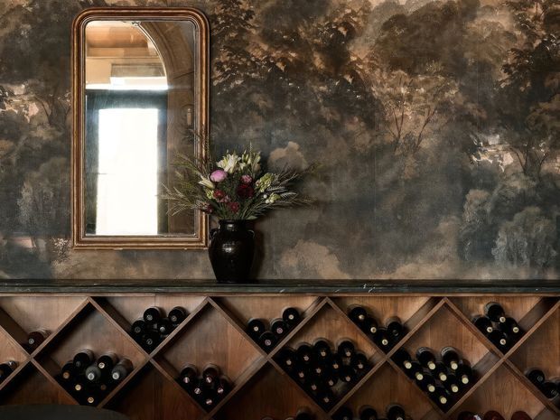 A richly styled wine storage cabinet or bar console occupies the lower half of the image, with diagonal wooden cubbies filled with wine bottles. Above it is a dark, atmospheric mural-style wall covering depicting trees or a wooded landscape. A tall antique-style framed mirror, floral arrangement, and black vase sit on the counter, creating a moody, heritage-inspired wine room or home bar setting.