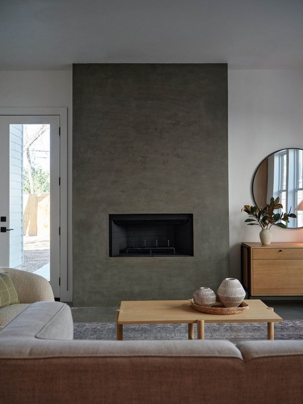 A calm, contemporary living room centered on a large dark grey textured fireplace surround with a recessed black fireplace. The room includes a neutral upholstered sofa in the foreground, a simple light timber coffee table, a mid-century-inspired timber sideboard, a round wall mirror, and a glazed exterior door. Soft natural light and muted neutral finishes create a refined, inviting atmosphere.