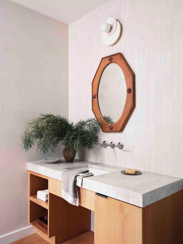 A stylish bathroom vanity featuring a long pale pink-white tiled wall, two wall-mounted basin mixers, light grey stone-look benchtops, and warm timber open shelving and cabinetry. A sculptural white wall light, octagonal timber-framed mirror, greenery, folded towels, and a draped hand towel add decorative detail. The space has a softly lit, warm contemporary character.