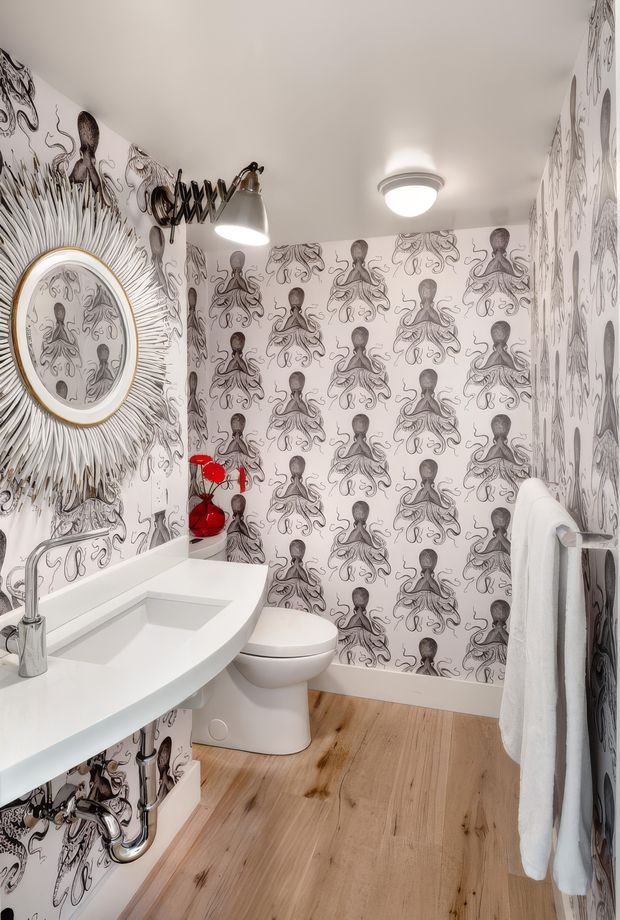 Compact eclectic bathroom with a white sculptural vanity and integrated basin, wall-mounted chrome plumbing, toilet, and patterned octopus wallpaper covering the walls. The room has warm timber flooring, a decorative sunburst mirror, black-and-brass wall lighting, a red accent object, and a white towel rail. Bright, playful, highly decorative interior.
