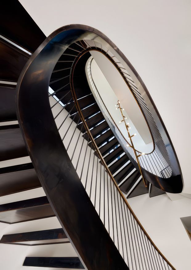 Dramatic upward view of a sculptural, multi-level staircase with a sweeping curved balustrade, dark metal or timber stair treads, slender vertical metal spindles, and warm-toned handrails. A bright open stairwell and linear pendant lighting emphasize the tall internal void and elegant spiral-like geometry.