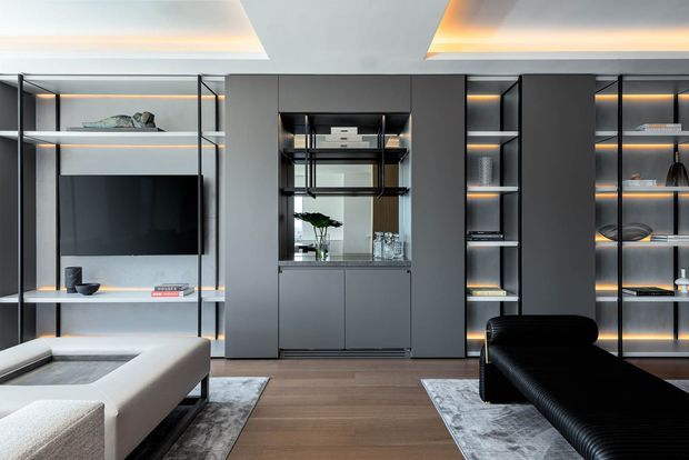 A sophisticated contemporary living room featuring a full-height built-in media and display wall in dark grey cabinetry, open shelving with integrated warm LED lighting, a central glazed bar or display niche, and a wall-mounted television. The room has timber flooring, pale area rugs, a large light-coloured sectional sofa, and a black leather lounge chair. Recessed ceiling details with concealed lighting add a refined, dramatic atmosphere.