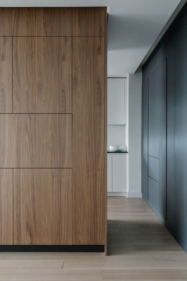 A refined minimalist interior corridor featuring full-height walnut-toned timber cabinetry or wall panelling on the left and a dark charcoal-blue paneled built-in wall on the right. Pale timber flooring leads toward a compact white joinery area with an integrated dark benchtop and a small basin or decorative vessel. The space has clean flush detailing, concealed storage, restrained colours, and soft natural light.