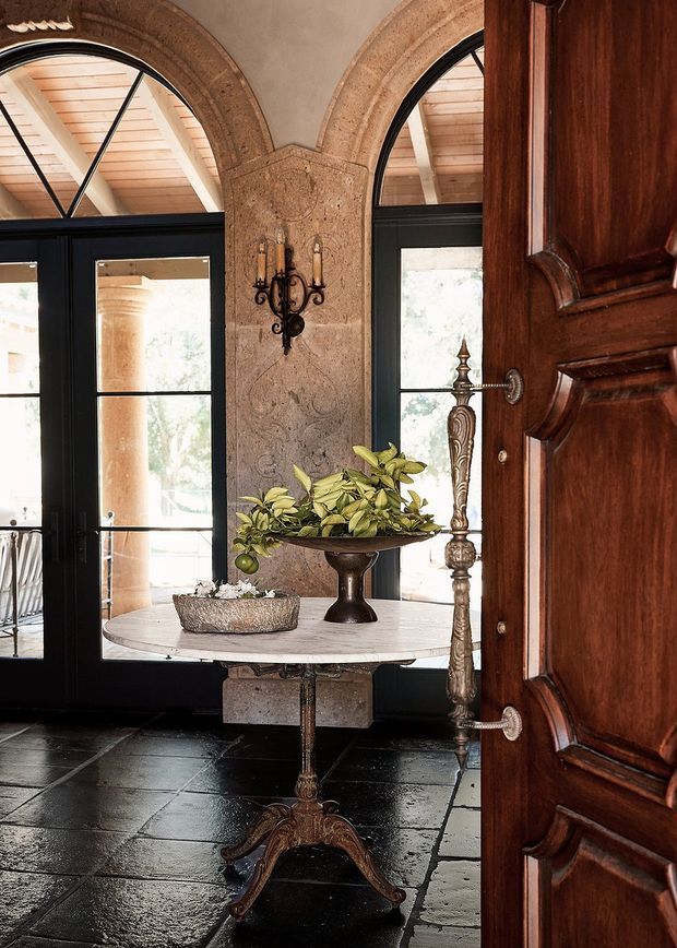 An ornate, traditional entry hall viewed through a partially open carved timber door. The space features arched black-framed glazed doors and windows, a textured stone or plaster arch surround, dark stone floor tiles, a round pedestal table with a decorative planter, and an elaborate wall sconce. Warm natural light enters through the glazing, highlighting the richly detailed architectural finishes.