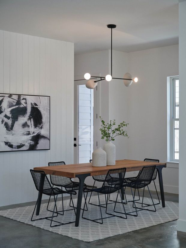 A bright, contemporary dining room with a substantial rectangular timber dining table surrounded by black woven wire-frame chairs. The room features white vertical wall panelling, pale walls, polished concrete flooring, a light patterned area rug, an abstract monochrome artwork, and a sculptural black multi-arm pendant light. A simple branch arrangement in white ceramic vessels sits at the centre of the table.