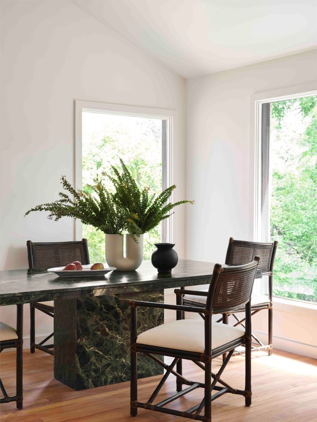 Bright dining area with a substantial dark green veined stone dining table, surrounded by woven cane-back wooden chairs. White walls, large white-framed windows, and light timber flooring create an airy setting with leafy greenery visible outside. A simple floral arrangement and black decorative vessel sit on the table.