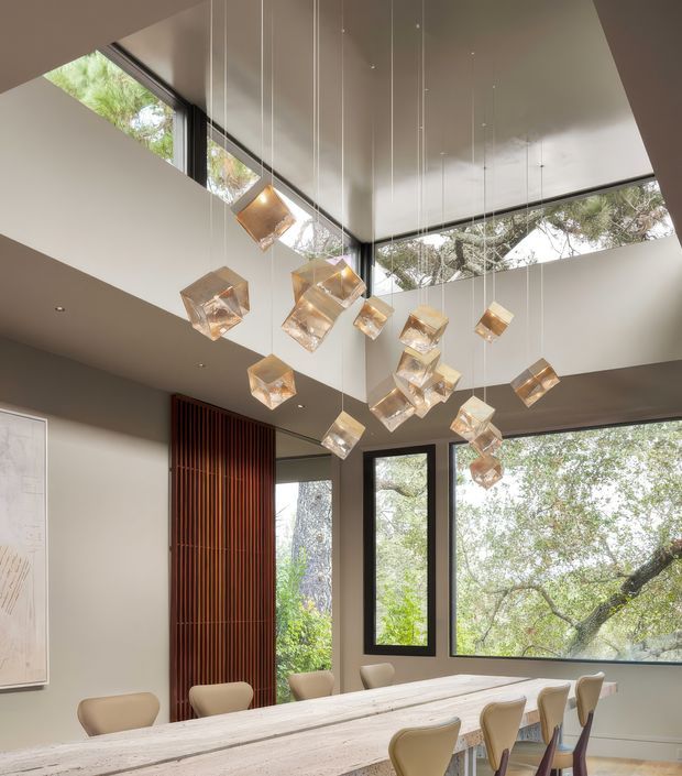 A contemporary dining room with a long pale timber dining table and upholstered dining chairs, framed by large black-framed windows overlooking mature trees. The room features a dramatic high ceiling with clerestory glazing and a sculptural cluster of suspended translucent pendant lights. A tall dark timber vertical screen forms a feature wall element beside the windows.