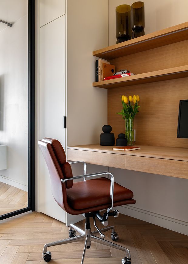 A compact built-in home office nook with warm timber shelving, a floating desk, and integrated cabinetry. A brown leather swivel office chair sits on a light timber herringbone floor beside a full-height glass door or partition. Decorative black vases, books, a small model car, and yellow tulips style the open shelving and desk area. The scheme is warm, refined, and contemporary.