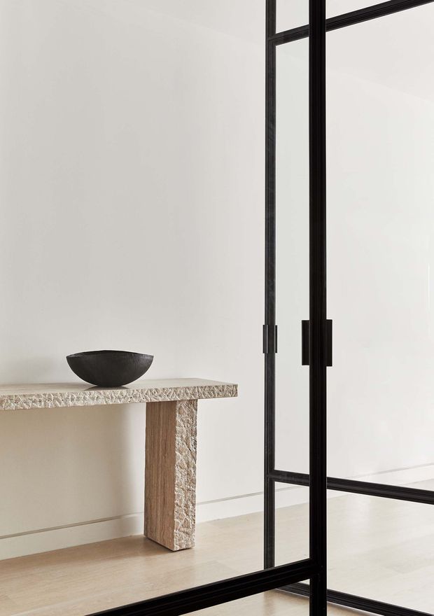 Minimalist interior vignette featuring a slim console table with a textured light stone top and substantial natural timber support, paired with a dark sculptural bowl. A tall black-framed glazed partition or internal door dominates the right side, with white walls and pale timber flooring creating a quiet, refined composition.