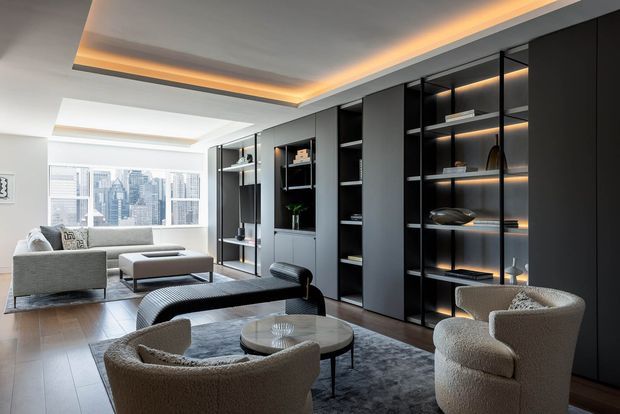 A sophisticated open-plan living room with a lounge area arranged around low round coffee tables and upholstered armchairs, plus a grey sofa near floor-to-ceiling glazing. A full-height dark built-in media and display wall with illuminated open shelving dominates one side of the room. The space has pale timber flooring, a large grey rug, a layered recessed ceiling with warm LED cove lighting, and strong urban skyline views through the windows.