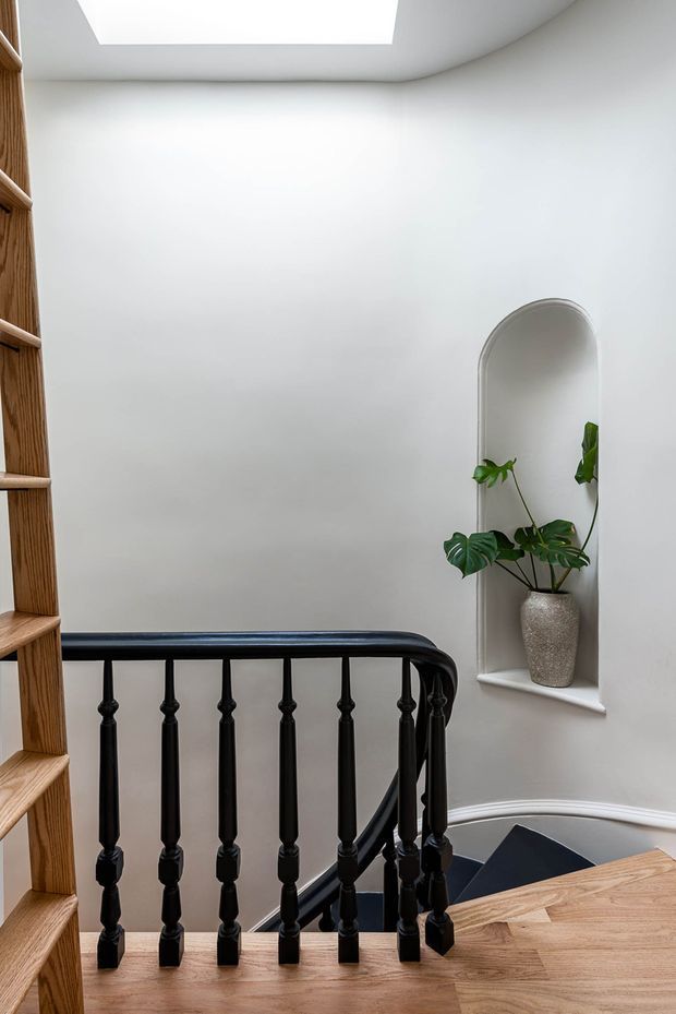 A bright residential stair landing with a traditional black turned-baluster railing, warm timber stairs and open shelving, white curved plaster walls, a skylight, and an arched recessed niche displaying a potted tropical plant.
