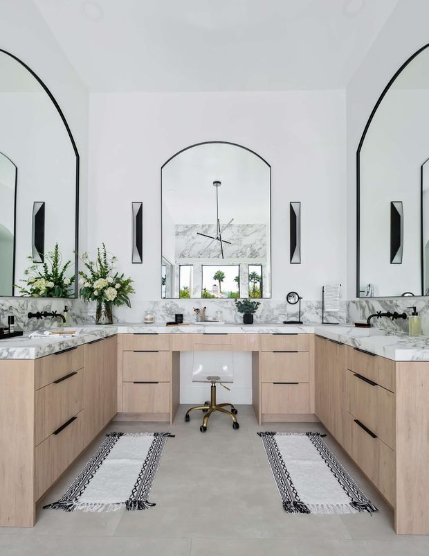 Bright, spacious bathroom vanity area with a symmetrical U-shaped light timber built-in cabinet arrangement, white marble-look benchtops and backsplash, large arched mirrors, black wall sconces and tapware, and a central seated makeup or dressing station. Two patterned runners sit on a pale stone-look floor. The room has a refined, contemporary classic appearance with abundant natural light.