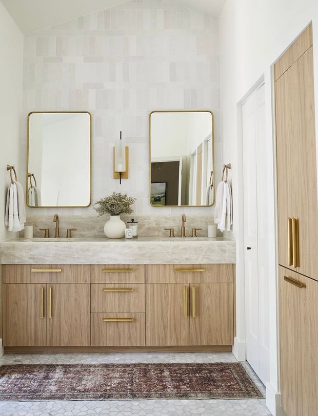 Bright, refined bathroom vanity with a double-sink timber cabinetry unit, pale stone benchtop, brass tapware and hardware, twin rounded-rectangle mirrors, white patterned wall tile, and hexagonal floor tile. A large rug and adjacent timber built-in cabinetry add warmth. The room has a high vaulted ceiling and a calm contemporary-traditional aesthetic.