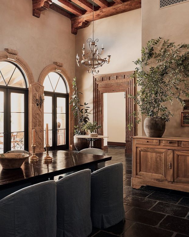 A richly detailed formal dining room with a dark wood dining table and upholstered chairs, large arched glazed doors framed by carved stone or plaster surrounds, an ornate carved timber mirror, timber cabinetry, a traditional chandelier, exposed timber ceiling beams, and dark stone flooring. The palette is warm and earthy with Mediterranean-inspired architectural detailing.