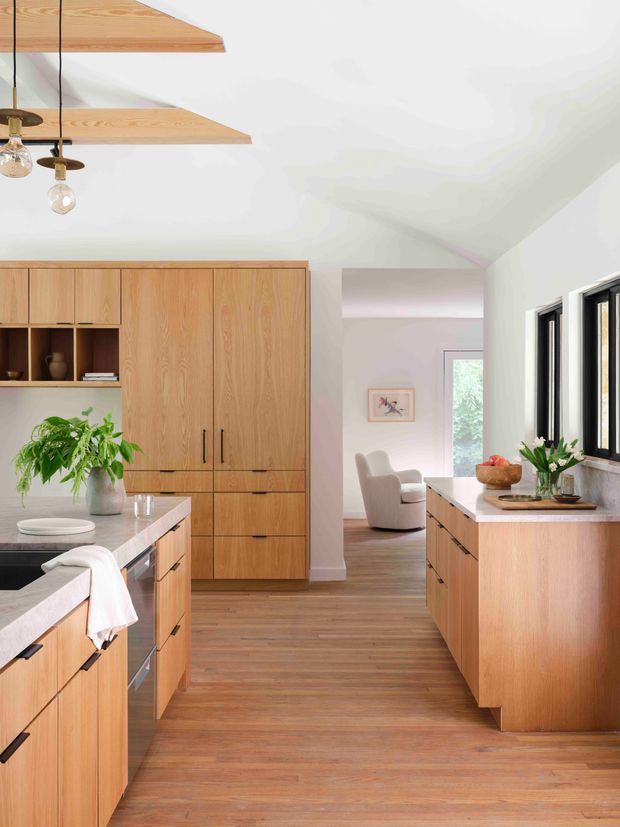 Bright contemporary kitchen with extensive warm timber cabinetry, pale stone benchtops, and a matching timber floor. The kitchen includes a central island, tall integrated storage, open shelving, and a secondary run of cabinetry beneath black-framed windows. Exposed timber ceiling beams and suspended glass pendant lights add character. A living area with an armchair is visible beyond the kitchen.