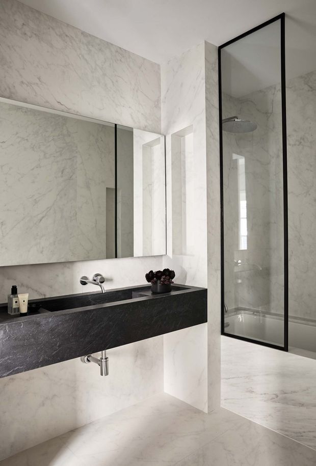 A refined contemporary bathroom clad extensively in white veined marble-look stone. The room features a wall-mounted dark stone vanity with an integrated basin, polished chrome wall-mounted tapware, a large frameless mirror, and a black-framed glass shower enclosure with a rainfall shower over a freestanding or partially visible bath. The palette is predominantly white and grey with strong black accents, creating a luxurious, minimalist atmosphere.