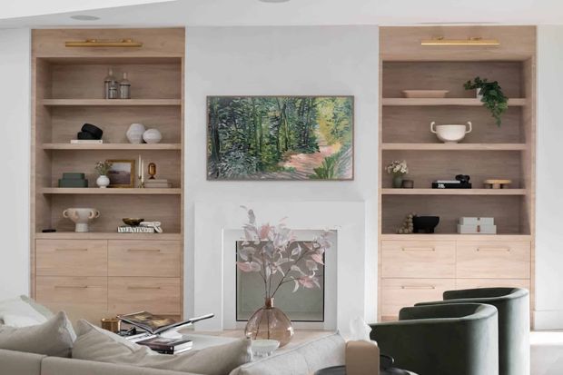 A refined living room with symmetrical built-in timber shelving and lower cabinetry flanking a central painted wall and artwork. The shelving displays ceramics, books, decorative objects and plants, while a pale sectional sofa and dark green curved armchairs occupy the foreground. Recessed ceiling lighting and warm neutral finishes create a calm, contemporary interior.