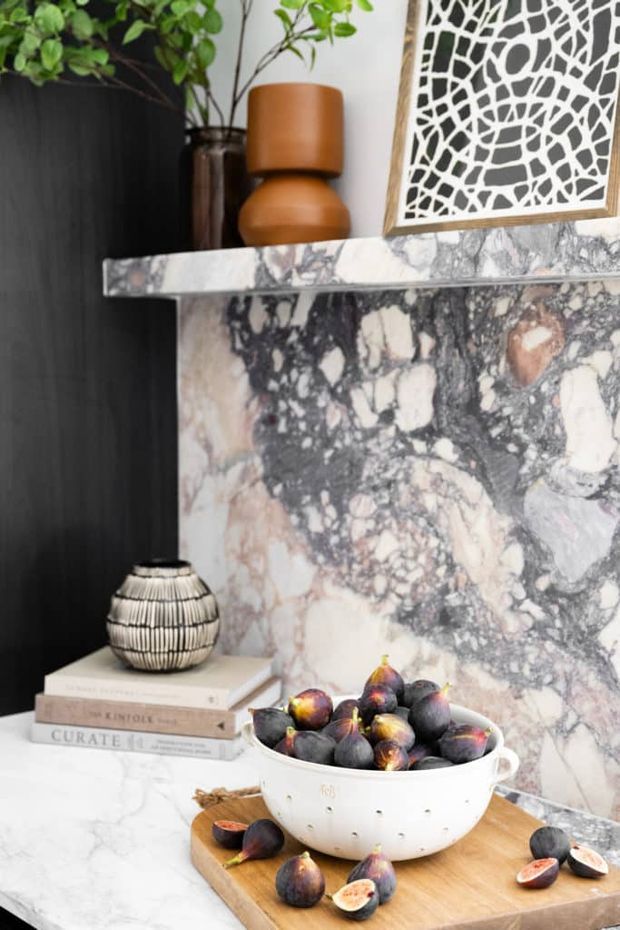 Decorative interior vignette featuring a dramatic grey, white and blush-veined marble fireplace surround and mantel. A dark vertical wall panel forms the backdrop, with a potted leafy plant, sculptural terracotta vessel, framed abstract artwork, and a ceramic bowl of figs on a timber board arranged on the marble surface. The image emphasizes the stone feature and layered styling.