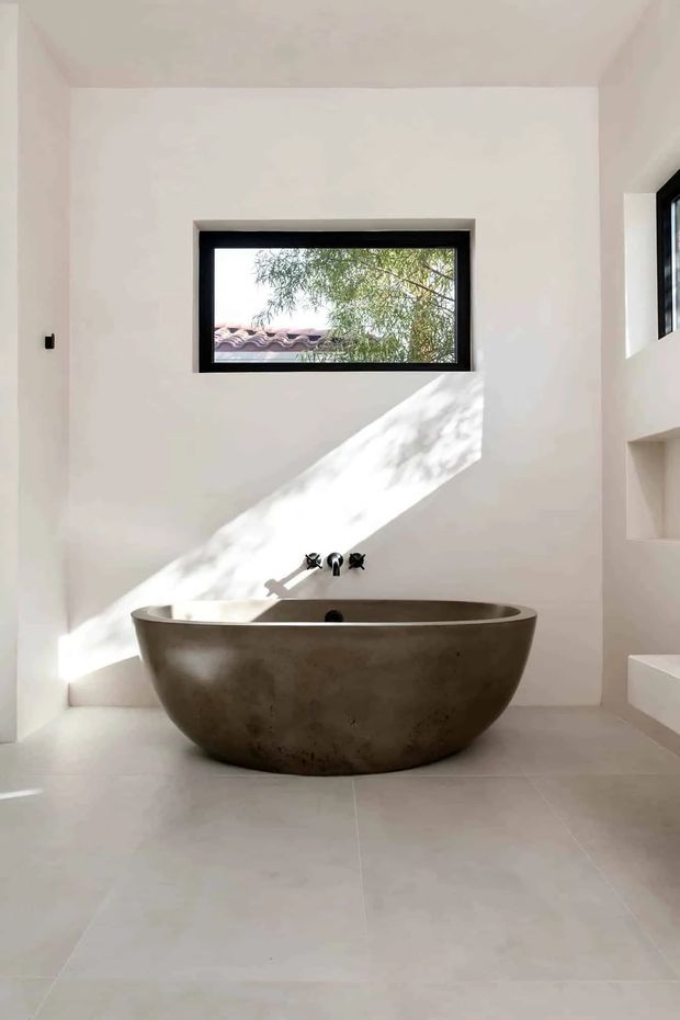 Bright minimalist bathroom featuring a large freestanding oval bathtub in a mottled dark stone or concrete finish. White plaster walls, pale large-format floor tiles, black-framed horizontal window, recessed shelving, and understated black wall-mounted bath fittings create a clean, sculptural interior. Strong diagonal sunlight and tree shadows add contrast and warmth.