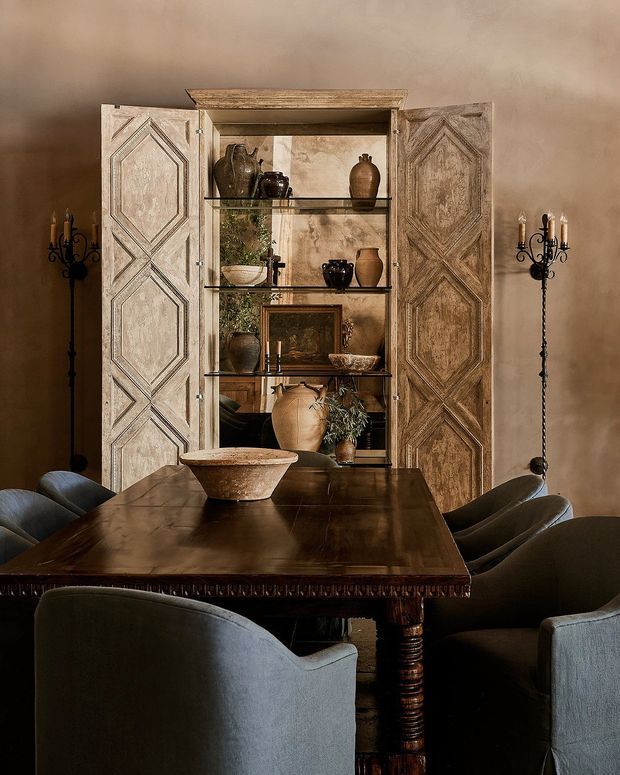 Moody, warmly lit dining room featuring a large dark timber dining table surrounded by upholstered dining chairs. A distressed carved timber display cabinet with open shelving occupies the rear wall, styled with pottery, greenery, framed art and decorative objects. Tall black wrought-iron candle stands flank the cabinet, while the walls have a warm textured plaster finish. The room has a richly layered, rustic traditional character.