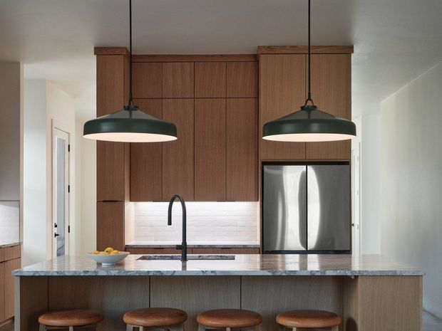Contemporary kitchen viewed from the living side, centered on a large island with four timber stools. The kitchen features full-height warm timber cabinetry, a dark stone-look benchtop, integrated stainless-steel refrigerator, black gooseneck tap, white horizontal-tile splashback, and two oversized black pendant lights. The space is bright and clean with a restrained, warm material palette.