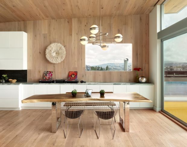 Warm open-plan kitchen and dining area enclosed in light timber panelling, with white built-in cabinetry, dark tiled splashback, pale timber flooring, and a substantial timber dining table with wire-frame chairs. Full-height glazing and a large window provide expansive views, while a sculptural multi-globe pendant light and textured wall decoration add visual interest.