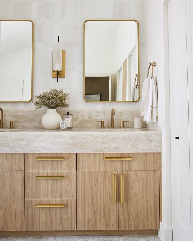 Bright, refined bathroom vanity with a double basin, pale veined stone benchtop and backsplash, vertically tiled wall, warm timber cabinetry, and brushed brass mirrors, tapware, handles, sconces, and towel hardware. Two rounded-rectangle mirrors sit above the vanity, with a white door visible at right and a patterned tile floor below.