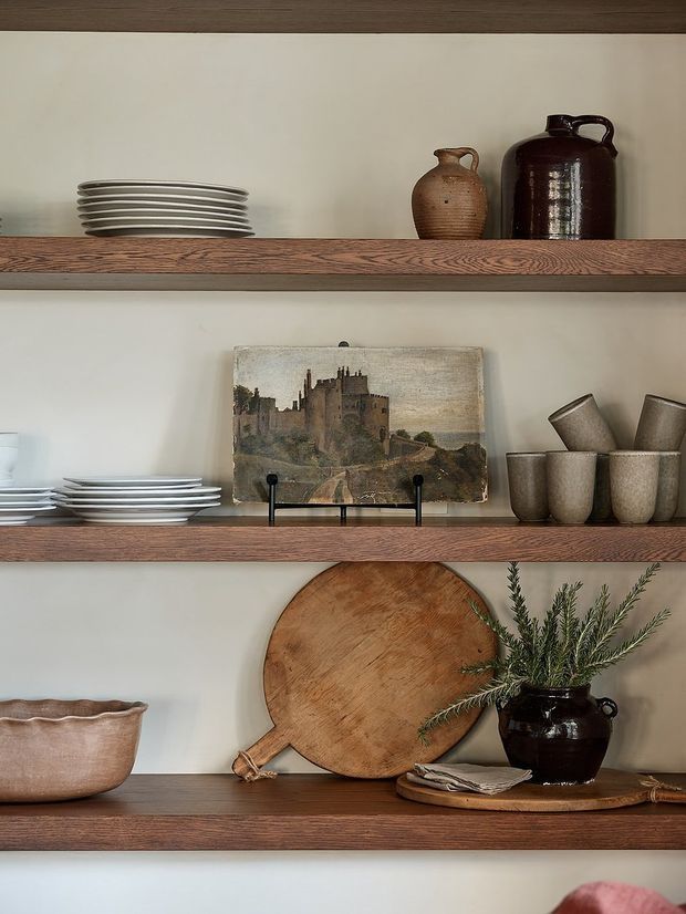 Close-up of three-tier open wooden shelving against a light plaster wall, styled with stacked ceramic plates, pottery vessels, metal cups, a framed castle artwork, a round wooden cutting board, a bowl, folded napkins, and a dark vase with greenery. The composition has a warm, rustic, curated display aesthetic.