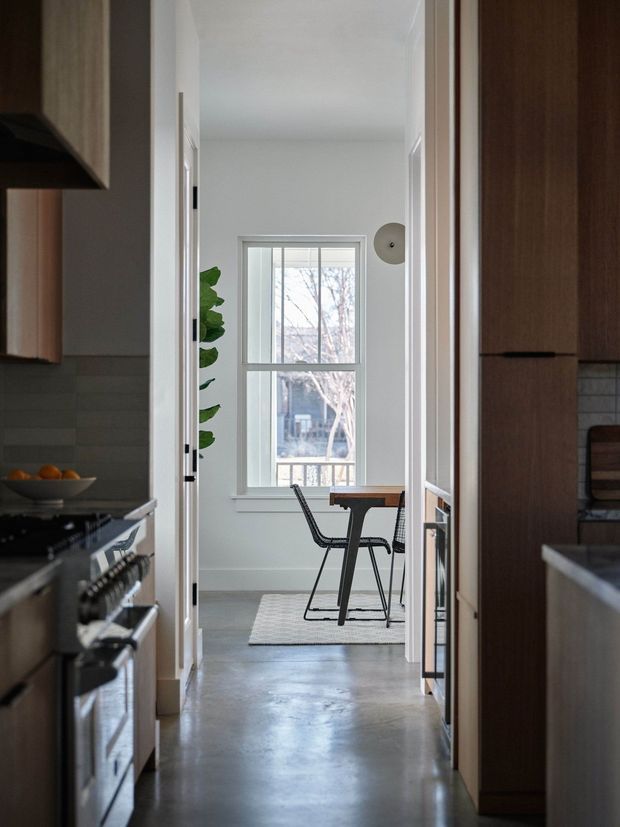 View down a narrow interior passage between a kitchen and a dining area. The foreground shows dark wood kitchen cabinetry, a stainless-steel oven and cooktop, and a muted tiled backsplash. At the far end, a small dining table with black wire-frame chairs sits beside a large white-framed window. The space has white walls, polished grey concrete flooring, simple contemporary detailing, and restrained natural light.