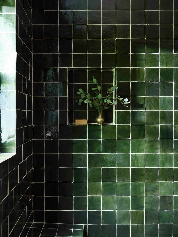 Close-up of a dark green glossy tiled wall corner, featuring a recessed tiled niche with a small brass vase and leafy branch. A narrow window or glazed opening is visible at the left, bringing in natural light. The image is dominated by the deep green tilework and reflective, moody finish.