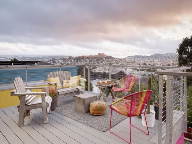 A spacious elevated outdoor deck or terrace furnished as a relaxed lounge and dining area, with composite or timber-look decking, a metal cable railing, weathered timber seating, colourful woven chairs, a small table, rug, planters, and broad elevated views over a densely built hilly urban landscape under a cloudy sky.