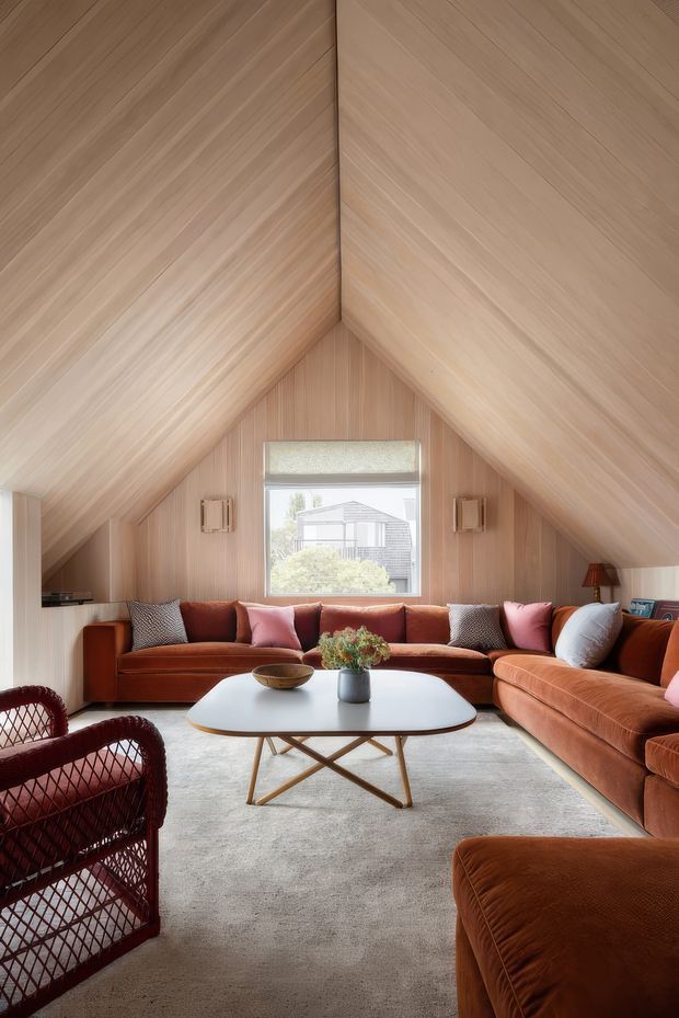 A warm, inviting living room beneath a steeply pitched, fully timber-lined vaulted ceiling. The room features rust and terracotta-toned sectional seating, woven lounge chairs, a pale rounded rectangular coffee table, a soft neutral carpet, and a central window with a roller blind. Vertical timber wall panelling and integrated wall lights create a cozy, refined cabin-like atmosphere.