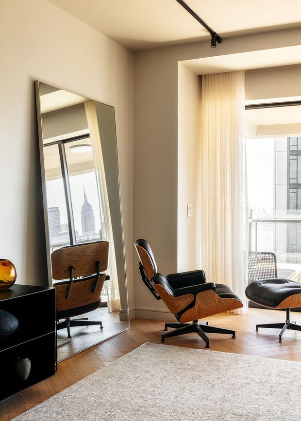 A refined contemporary living room with a mid-century-inspired lounge chair and matching ottoman positioned beside a large floor-to-ceiling window. Warm timber flooring, a large neutral rug, sheer curtains, a full-height leaning mirror, black low-profile cabinetry, and a linear black ceiling track light create a calm, warm interior. A city skyline is visible through the glazing and reflected in the mirror.