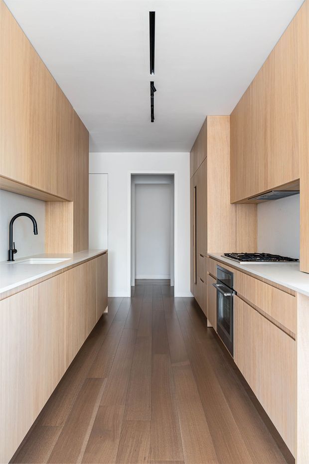 A narrow galley kitchen with full-height light timber cabinetry on both sides, white benchtops, a white ceiling and walls, dark timber plank flooring, black tapware, black linear ceiling lighting, and integrated cooking appliances. The kitchen opens to a hallway at the rear, creating a clean, streamlined contemporary interior.