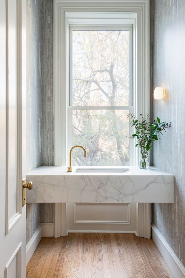 A compact, elegant powder room with a wall-mounted marble vanity beneath a tall white-trimmed window. The vanity includes an integrated rectangular basin and a brass gooseneck tap, while grey textured wallcovering, white panelled trim, warm timber flooring, and a small wall sconce create a refined traditional interior. A vase of greenery sits beside the basin.