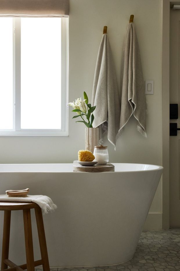 A calm, light-filled bathroom featuring a freestanding white bathtub beneath a window, two neutral towels hung on wooden pegs, and a small wooden stool with a folded towel and bath brush. A stone-like patterned floor, pale walls, and understated natural styling create a warm, serene atmosphere.
