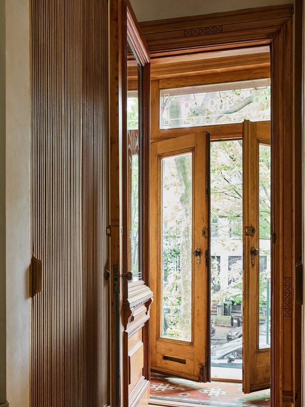 A tall, richly detailed timber-lined entryway featuring ornate carved trim, fluted wood wall panelling, glazed timber double doors, and patterned encaustic-style floor tiles. Natural light enters through the doors and transom, with greenery and an urban street visible outside.