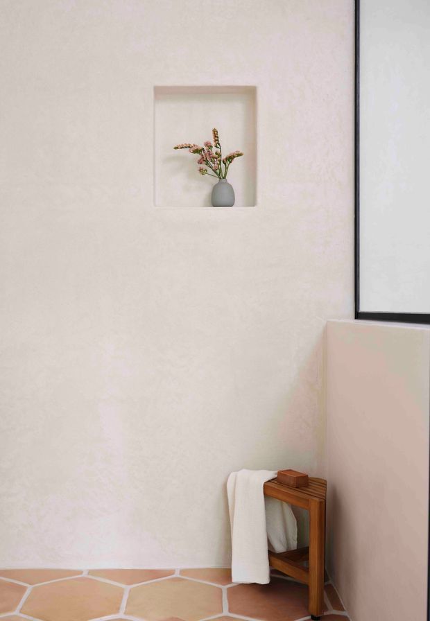 A minimalist bathroom corner with warm off-white textured walls, terracotta irregular polygon floor tiles with pale grout, and a black-framed window or shower glazing at right. A small recessed wall niche displays a simple grey vase with delicate flowers, while a compact wooden stool or bench holds folded white towels and a small wooden accessory. The composition is calm, warm, and understated.
