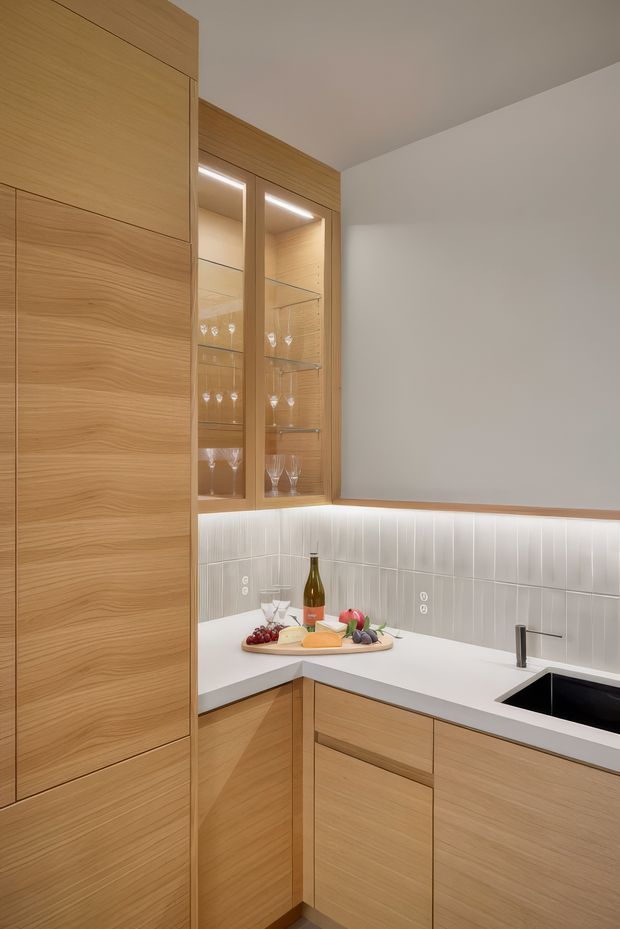 Compact contemporary kitchen or entertaining nook with full-height light timber cabinetry, a white L-shaped benchtop, integrated black sink and tap, and a vertically fluted white tiled splashback. Illuminated glass-front display cabinets contain wine glasses, while fruit, cheese and a wine bottle sit on the counter.
