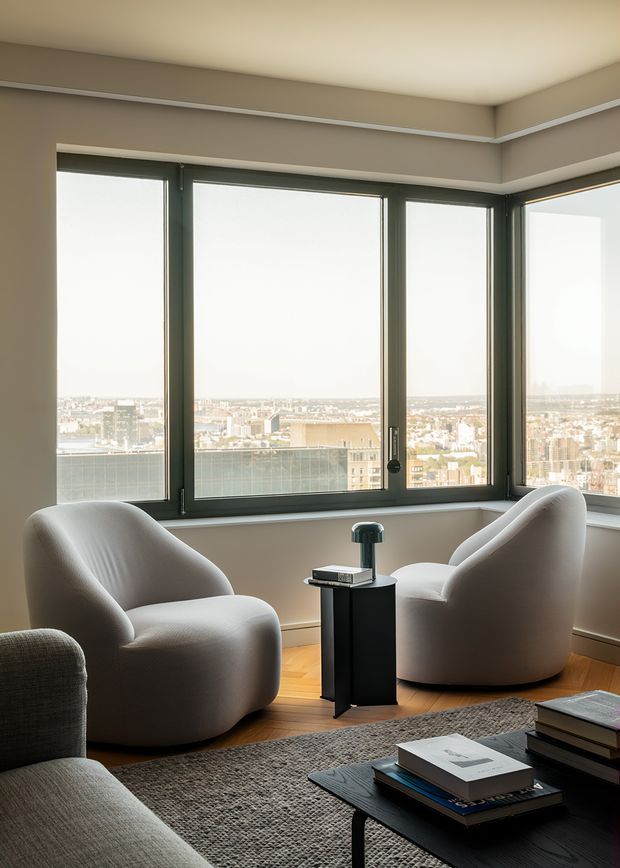 A refined high-rise living room with expansive corner windows overlooking a dense cityscape. Two sculptural cream lounge chairs sit beside a compact black pedestal table, with a grey sofa, textured rug, and low black coffee table with books in the foreground. Warm herringbone timber flooring, taupe walls, and dark window frames create a calm, contemporary setting with abundant natural light.