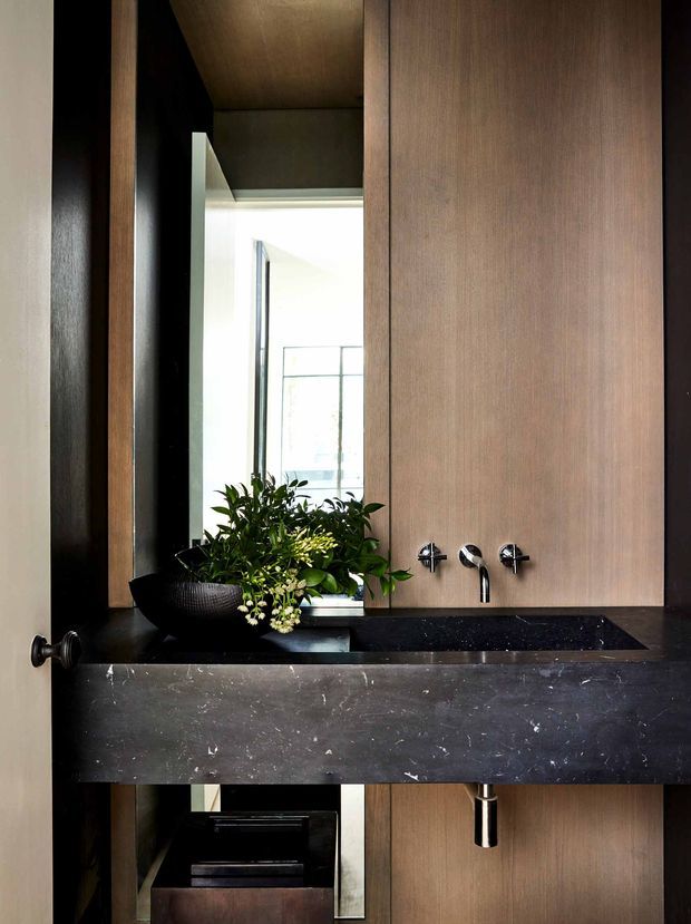 A compact, highly finished bathroom vanity viewed straight on. A dark stone benchtop with integrated rectangular basin spans the frame, paired with wall-mounted chrome tapware and warm vertical timber wall panelling. A tall mirror reflects a bright adjoining space, while a black vessel planter adds greenery. The composition is dramatic and refined, with strong contrast between dark stone, black accents, timber, and bright reflected light.