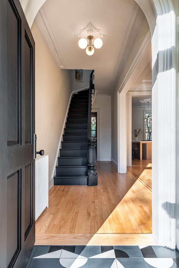 A bright, refined residential entry hall viewed from the front doorway. The space features a narrow black-painted staircase with turned newel posts, warm timber flooring, cream walls, ornate ceiling cornices and a decorative plaster ceiling medallion surrounding a three-globe pendant. A geometric black, white and grey tiled threshold contrasts with the timber floor. A doorway opens to a dining or kitchen area on the right, and tall sheer curtains frame the entry.