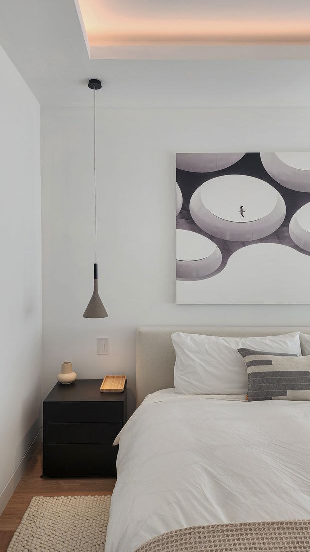 A calm, contemporary bedroom corner with a white upholstered bed, crisp white bedding, black bedside table, and a slim pendant light. A large abstract geometric artwork hangs above the bed. Warm timber flooring, a cream textured rug, soft neutral textiles, and a recessed ceiling detail with concealed warm lighting create a refined minimalist atmosphere.