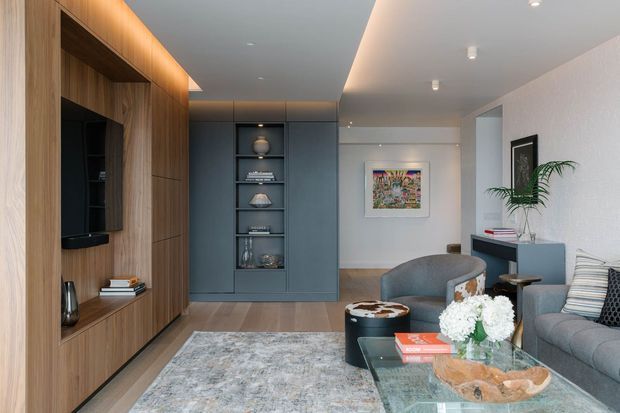 A refined contemporary living room with a warm timber feature wall and integrated media joinery on the left, a muted blue-grey built-in display cabinet at the rear, and grey upholstered seating arranged around a glass coffee table. Soft concealed and recessed lighting, a textured pale rug, artwork, and indoor plants create a calm, polished interior.