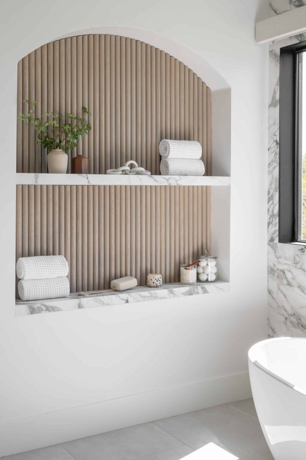 Bright, refined bathroom interior featuring a built-in recessed display niche with two arched openings, vertical fluted timber-toned backing, and white marble-look shelves. Folded white towels, small vessels, and toiletries are arranged in the niches. A marble-look wall surrounds a black-framed window, and the edge of a freestanding white bathtub is visible at lower right. The palette is predominantly white, light wood, and pale grey, with strong natural light.