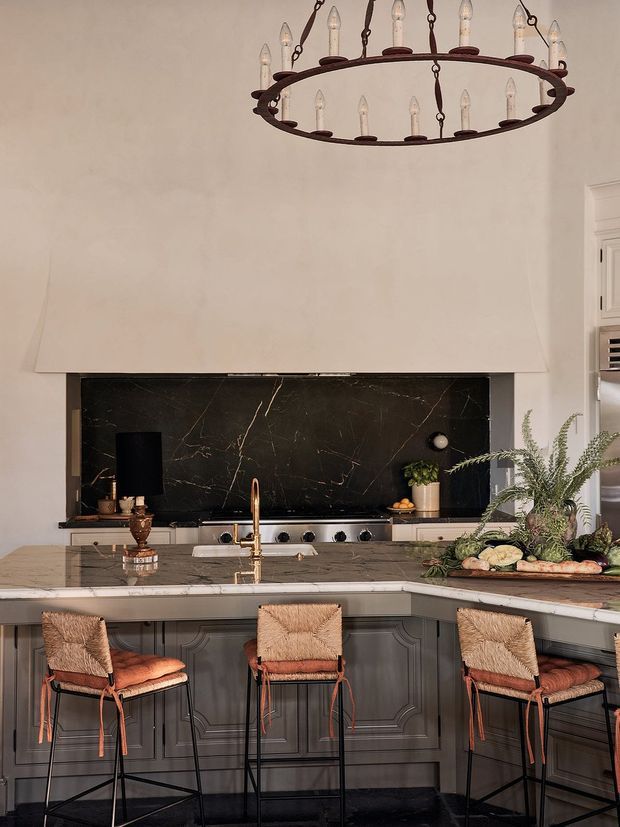 A richly styled kitchen with a large island and three woven bar stools. Dark grey decorative-panel cabinetry contrasts with a pale stone countertop, a dramatic black veined stone backsplash, brass tapware, and a large rustic ring chandelier. Fresh greenery and produce add an organic accent.