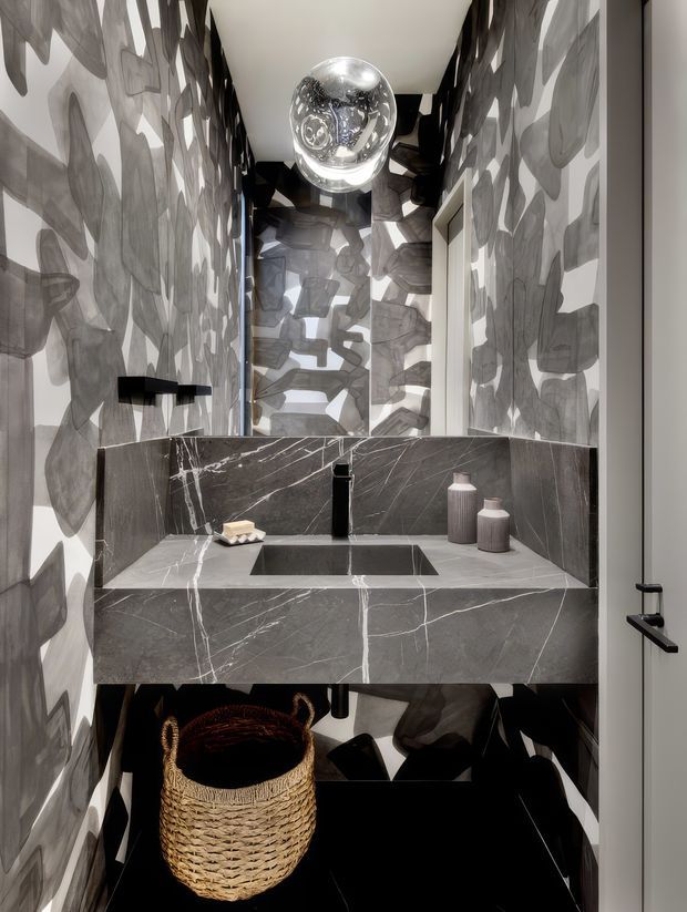 A compact, highly designed powder room with a floating dark marble vanity and integrated rectangular basin. The walls and ceiling are wrapped in a dramatic monochrome abstract pattern, complemented by a spherical pendant light, black tapware, a frameless mirror, and a woven basket beneath the vanity.