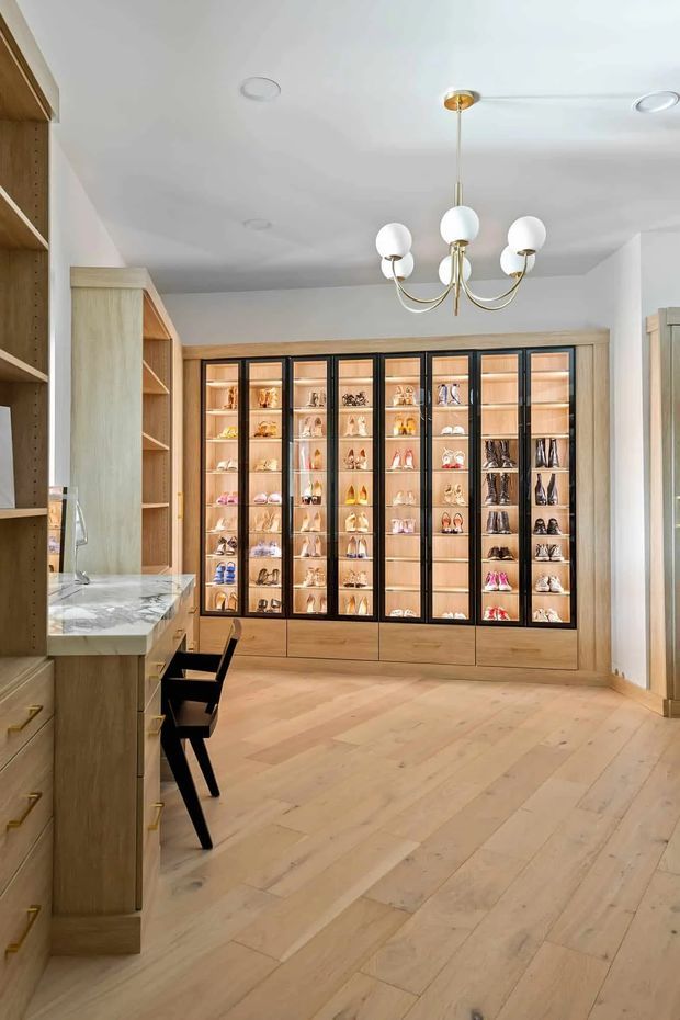 Bright, refined walk-in wardrobe or dressing room with extensive built-in light timber cabinetry, illuminated glass-front shoe and accessory display units, and a marble-look counter with a small black stool. The room has pale timber flooring, white walls and ceiling, brass-and-glass globe chandelier, and warm integrated cabinet lighting.