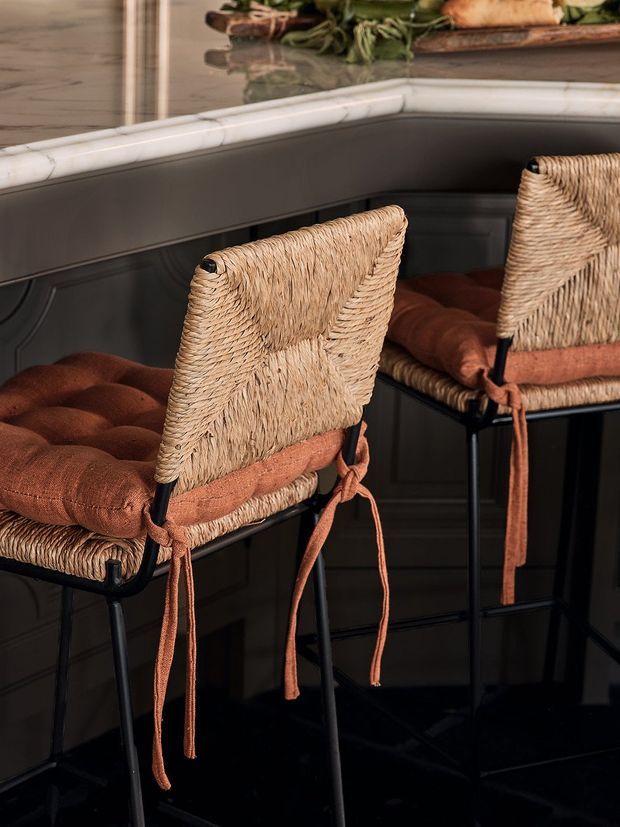 Close-up interior view of two woven rattan bar stools with rust-coloured tufted cushions and tie-on straps, positioned at a dark kitchen or bar counter. The space features dark panelled cabinetry, a pale veined stone benchtop, and glossy dark flooring, with a decorative shelf visible above.