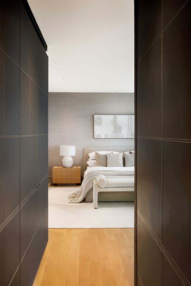 View through dark built-in cabinetry or a narrow passage into a refined bedroom. The room features a low upholstered bed with layered neutral bedding, a pale textured wall, minimalist abstract artwork, a warm timber bedside chest, and a sculptural white table lamp. Light timber flooring continues toward a large pale rug, while the dark joinery framing the view creates strong contrast.